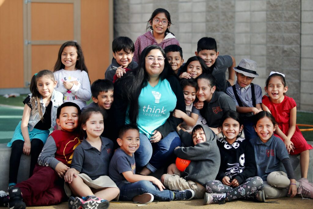 Teacher surrounded by a group of smiling elementary school children posing together outdoors.